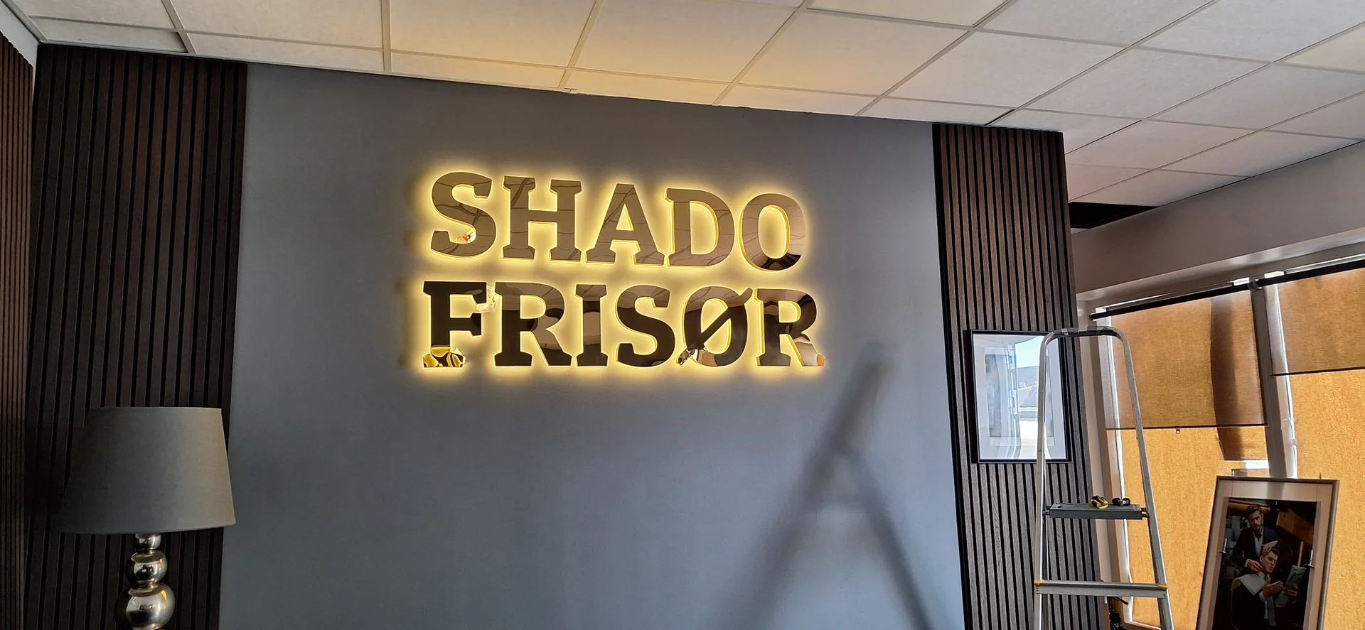 Gold SHADO FRISØR three-dimensional letters with warm backlighting on a dark interior wall