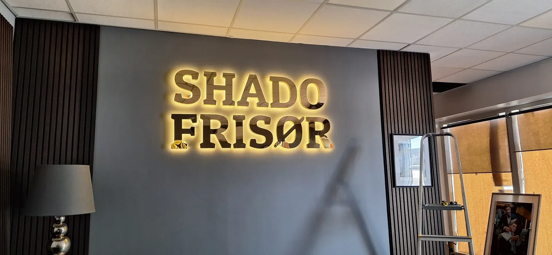 Gold SHADO FRISØR three-dimensional letters with warm backlighting on a dark interior wall