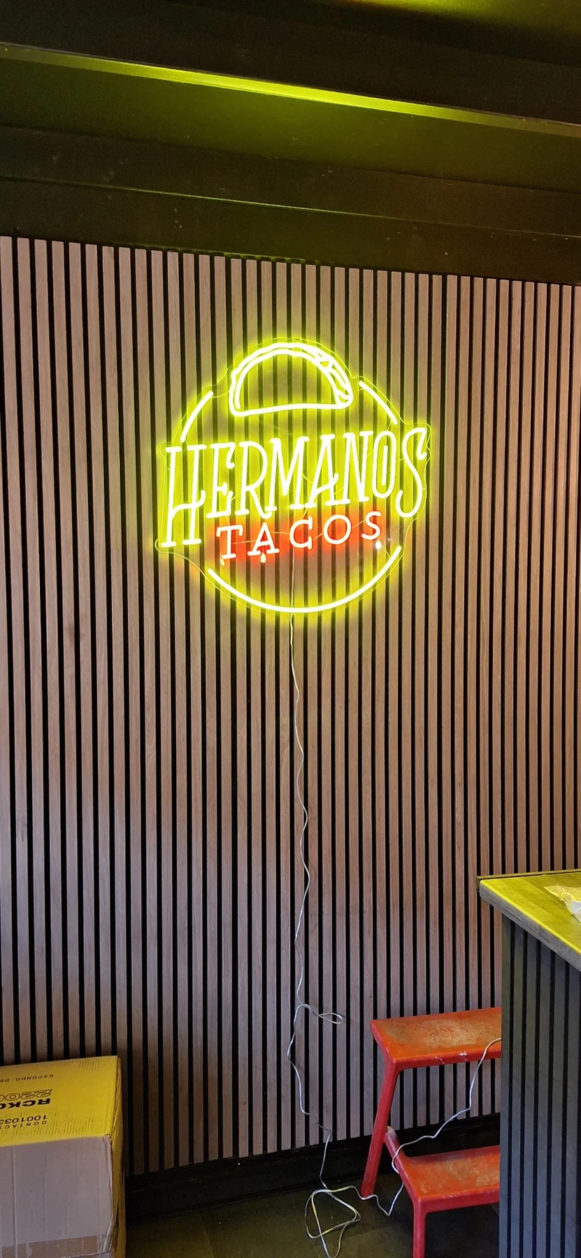 Illuminated HERMANOS TACOS LED logo on a vertical slatted wall